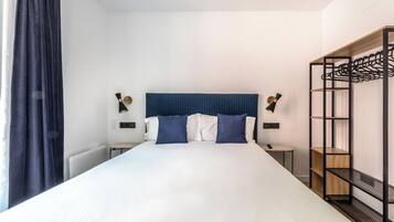 Una cama bien hecha con una decoración en blanco y azul, un cabecero azul oscuro y una estantería metálica negra a la derecha.