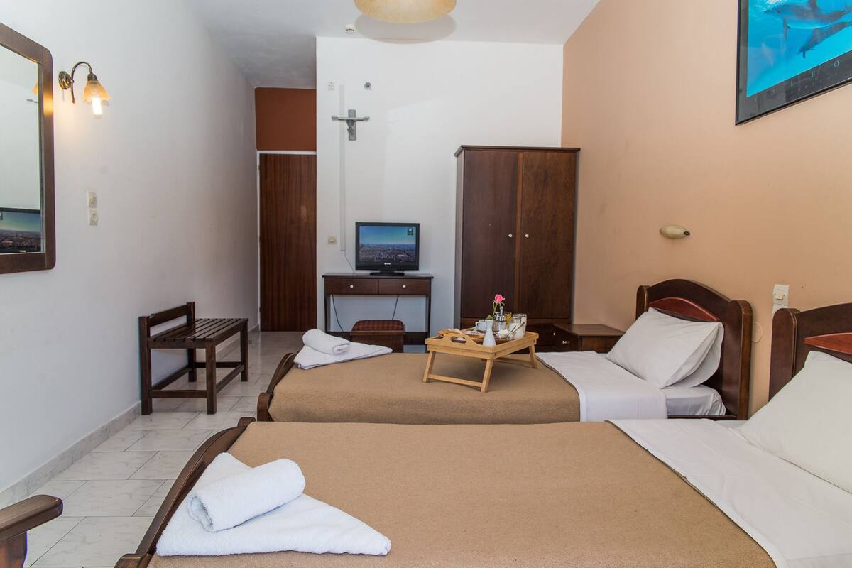 A hotel room with two beds, a desk with a computer, a wooden wardrobe, and a bench.