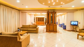 A spacious living area with a sofa, armchairs, a coffee table, and a television. There is a wooden cabinet and a television on a stand. The room has a tiled floor and a ceiling with recessed lighting.