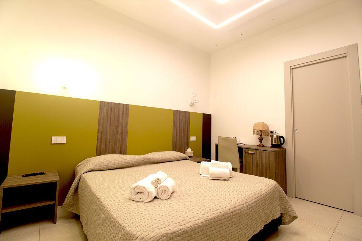 A hotel room with a bed, a nightstand, a lamp, a mirror, and a door.