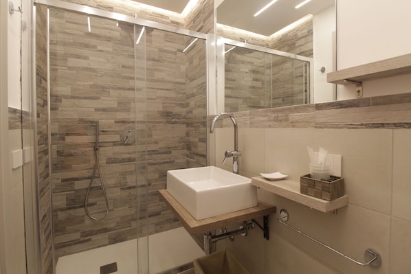 Un baño moderno con ducha de vidrio, un lavamanos blanco y una estantería de madera con artículos de aseo.