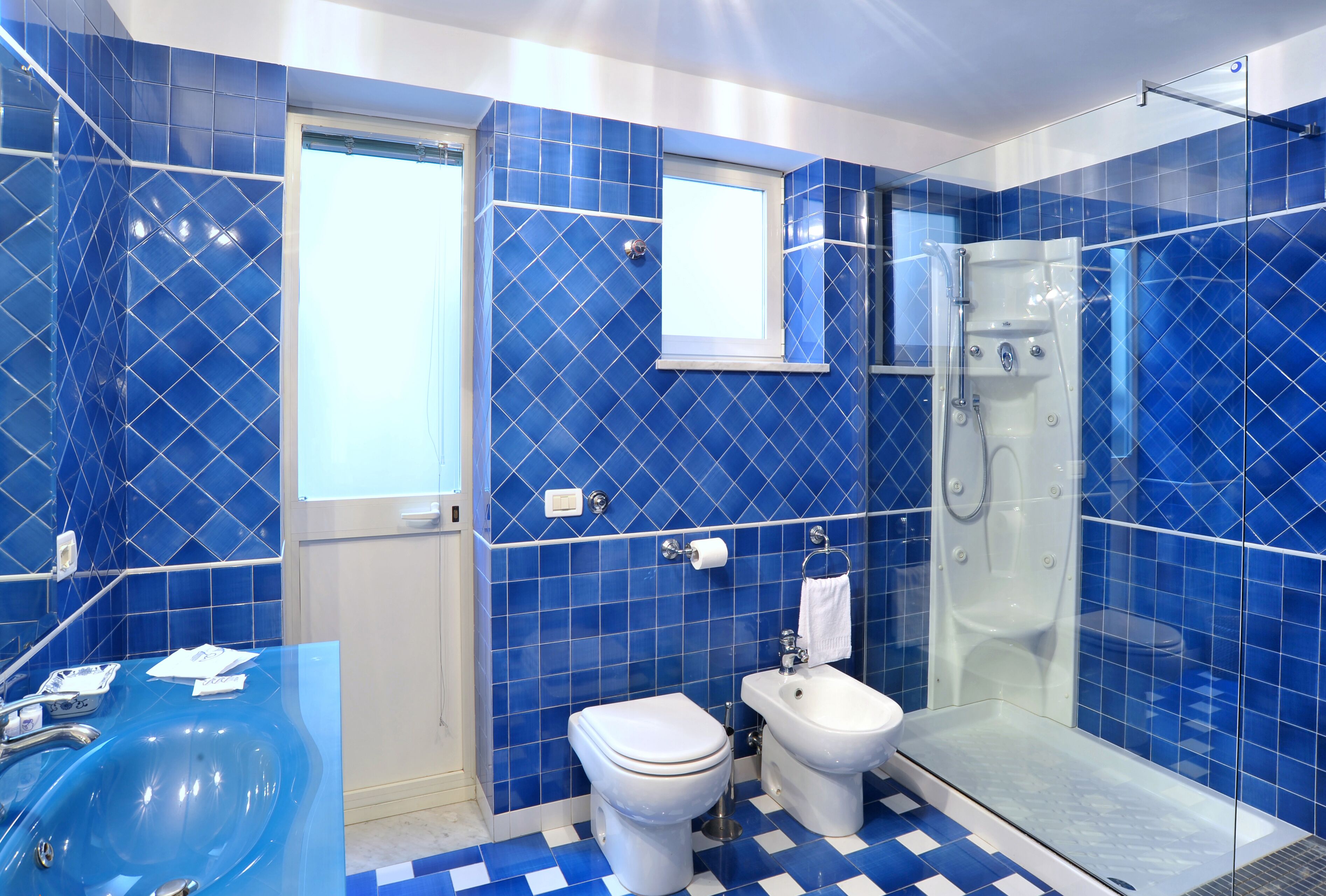 Un baño con paredes de azulejos azules, cabina de ducha con paredes de vidrio, inodoro blanco y lavamanos con encimera de vidrio.