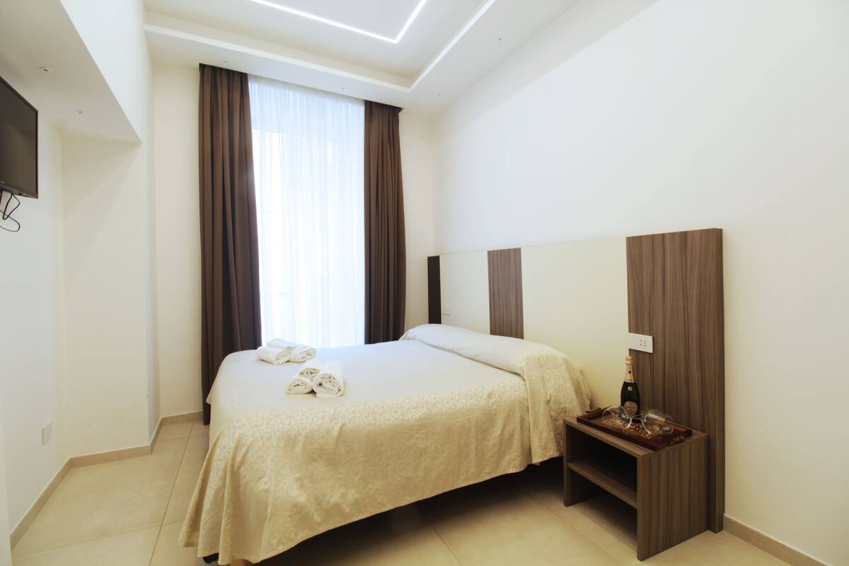 A hotel room with a bed, a bedside table with a bottle and glasses, a flat-screen TV mounted on the wall, and a window with curtains.