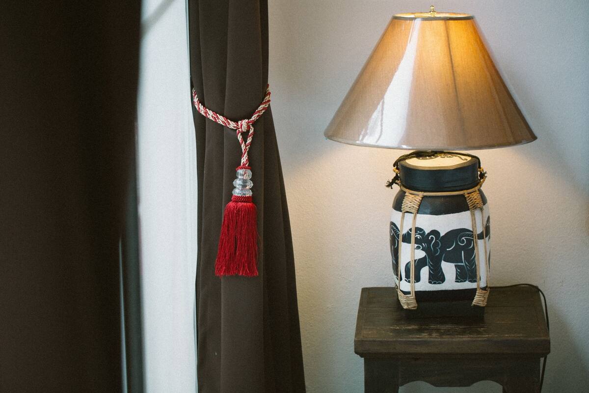 Una lámpara con pantalla cónica sobre una mesa de madera junto a una cortina con borlón.