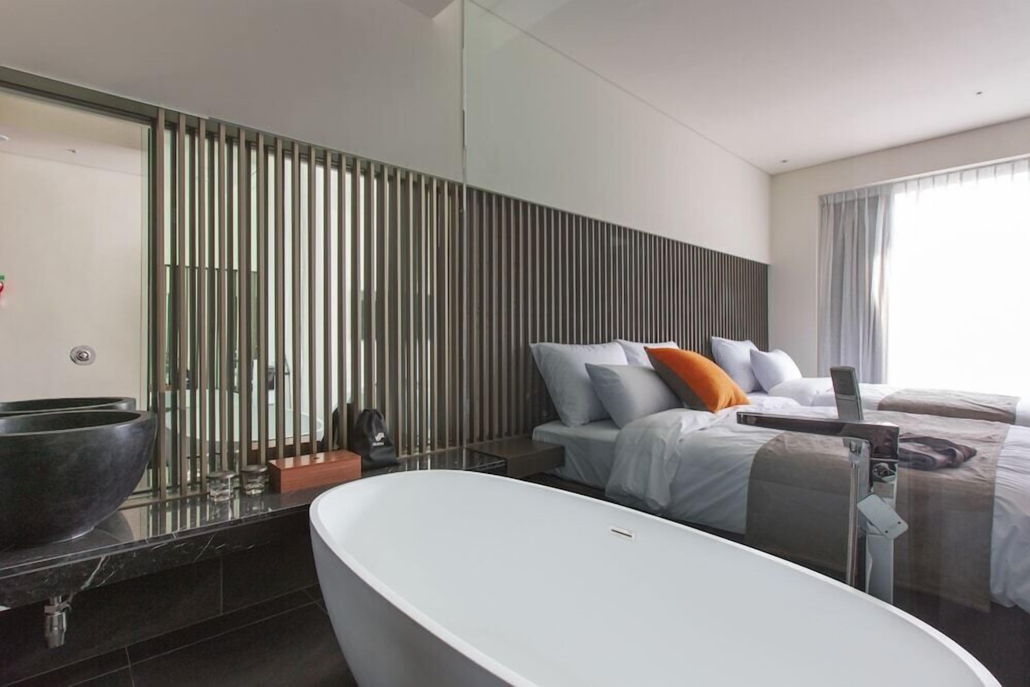 A modern hotel room with a large bathtub, a bed with pillows, a bedside table, and a large mirror.