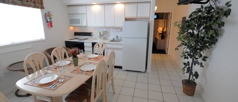A dining area with a table set for four, a kitchen in the background, and a potted plant on the right.