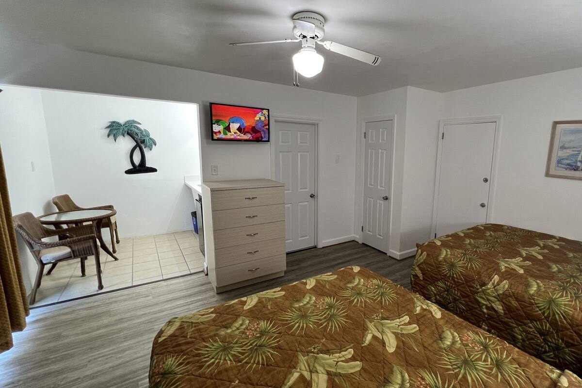 A room with two beds, a desk, a chair, a dresser, a ceiling fan, and a television.