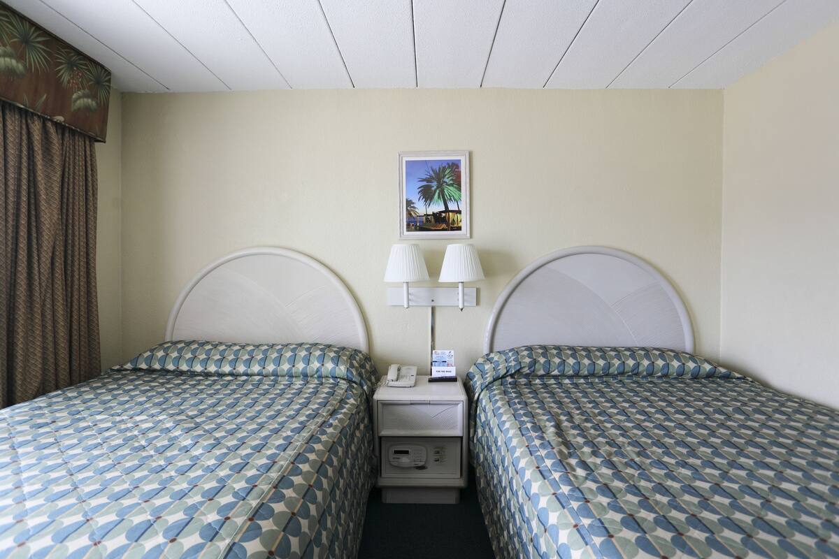 A hotel room with two beds, a nightstand, a lamp, a phone, and a picture on the wall.