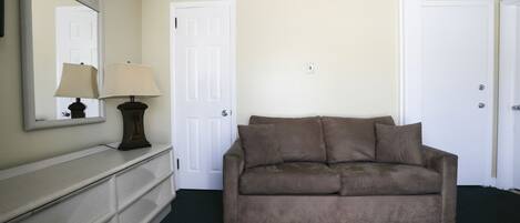 Una habitación con pared color beige, sofá marrón, puerta blanca y espejo con lámpara sobre un aparador.