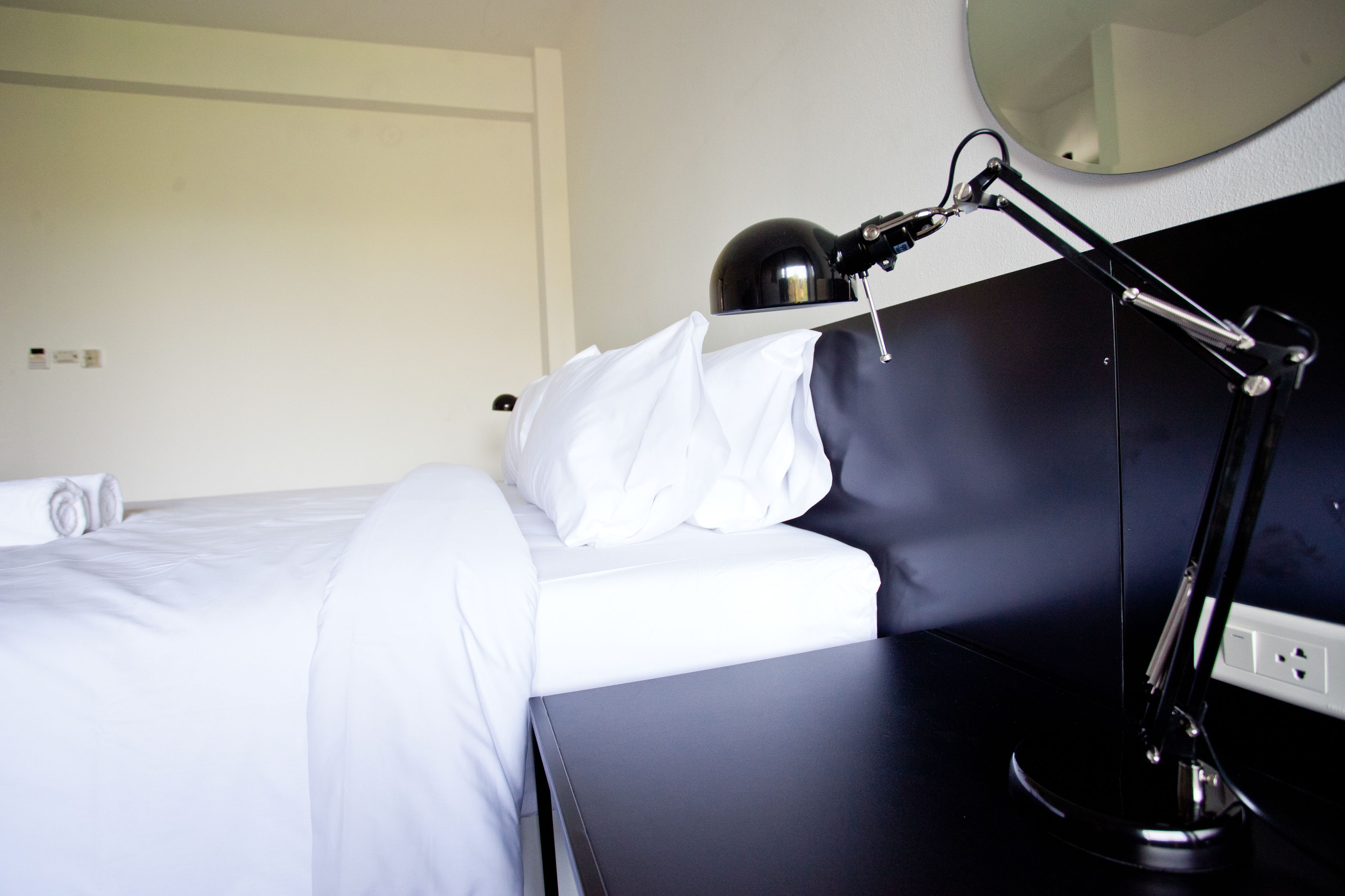 Una habitación de hotel con una cama bien hecha, una mesita de noche negra, una lámpara de escritorio y un espejo redondo en la pared.