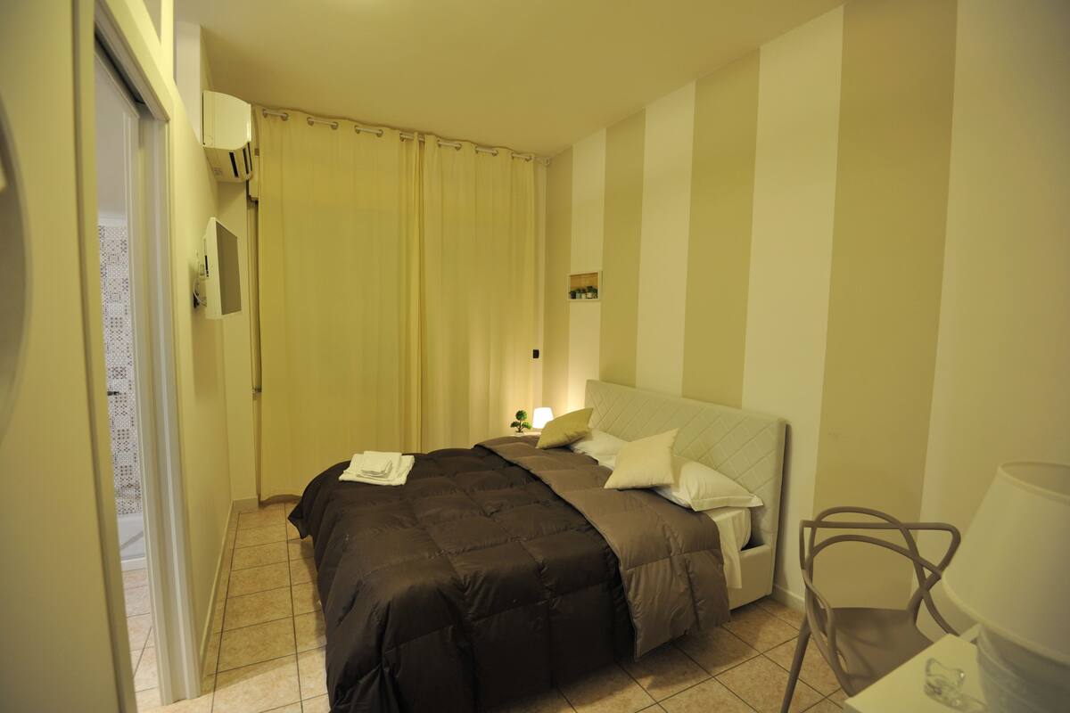 Quarto de hotel com cama, poltrona, mesa de cabeceira, luminária e cortina.