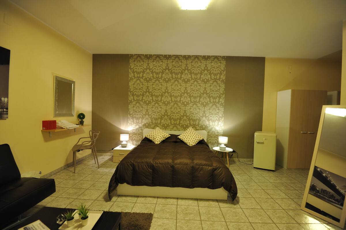 Quarto com cama grande, duas mesas de cabeceira com luminárias, um refrigerador pequeno, escrivaninha com cadeira e um espelho na parede.