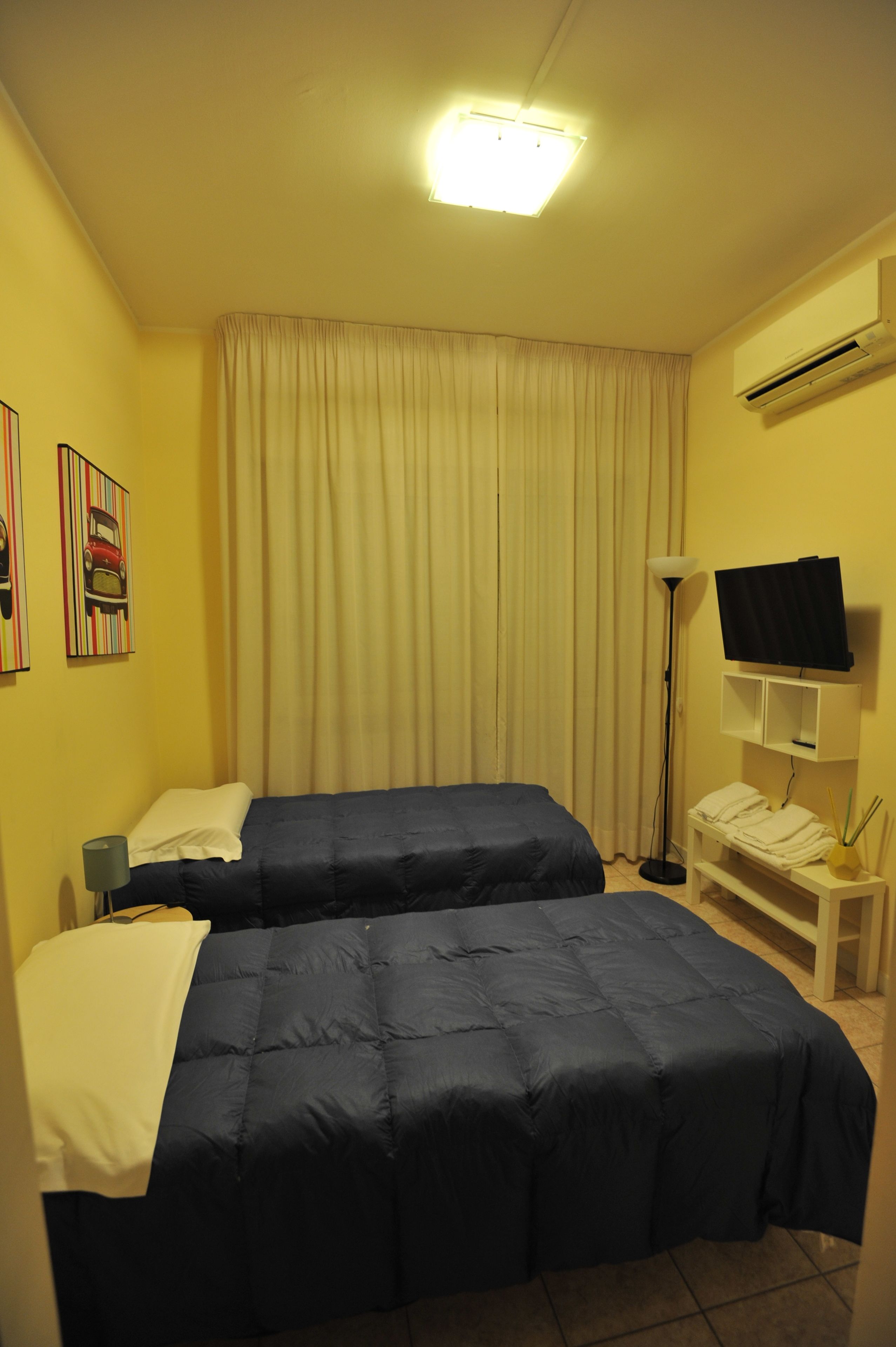 Habitación de hotel con dos camas, una lámpara de techo, televisión, mesita de noche con lámpara y ventana con cortinas.