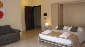 A hotel room with a bed, a bedside table with a lamp, a sofa, and a door leading to another room.