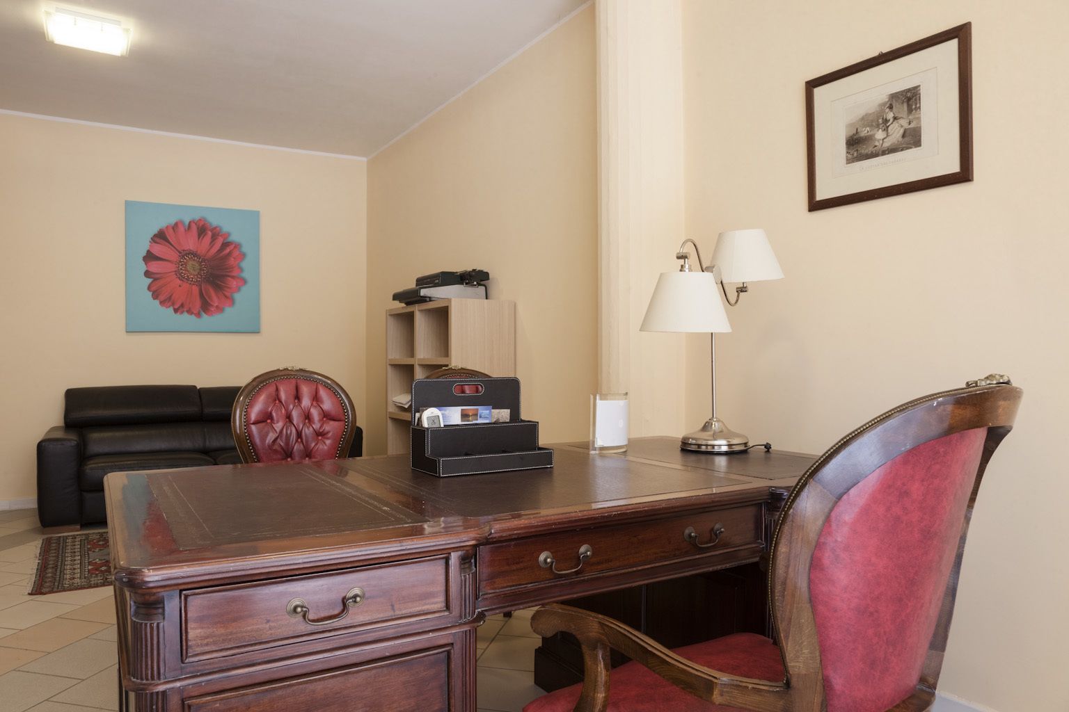 Habitación de hotel con un escritorio de madera, una silla roja, un sofá de cuero negro, un cuadro en la pared y una lámpara.