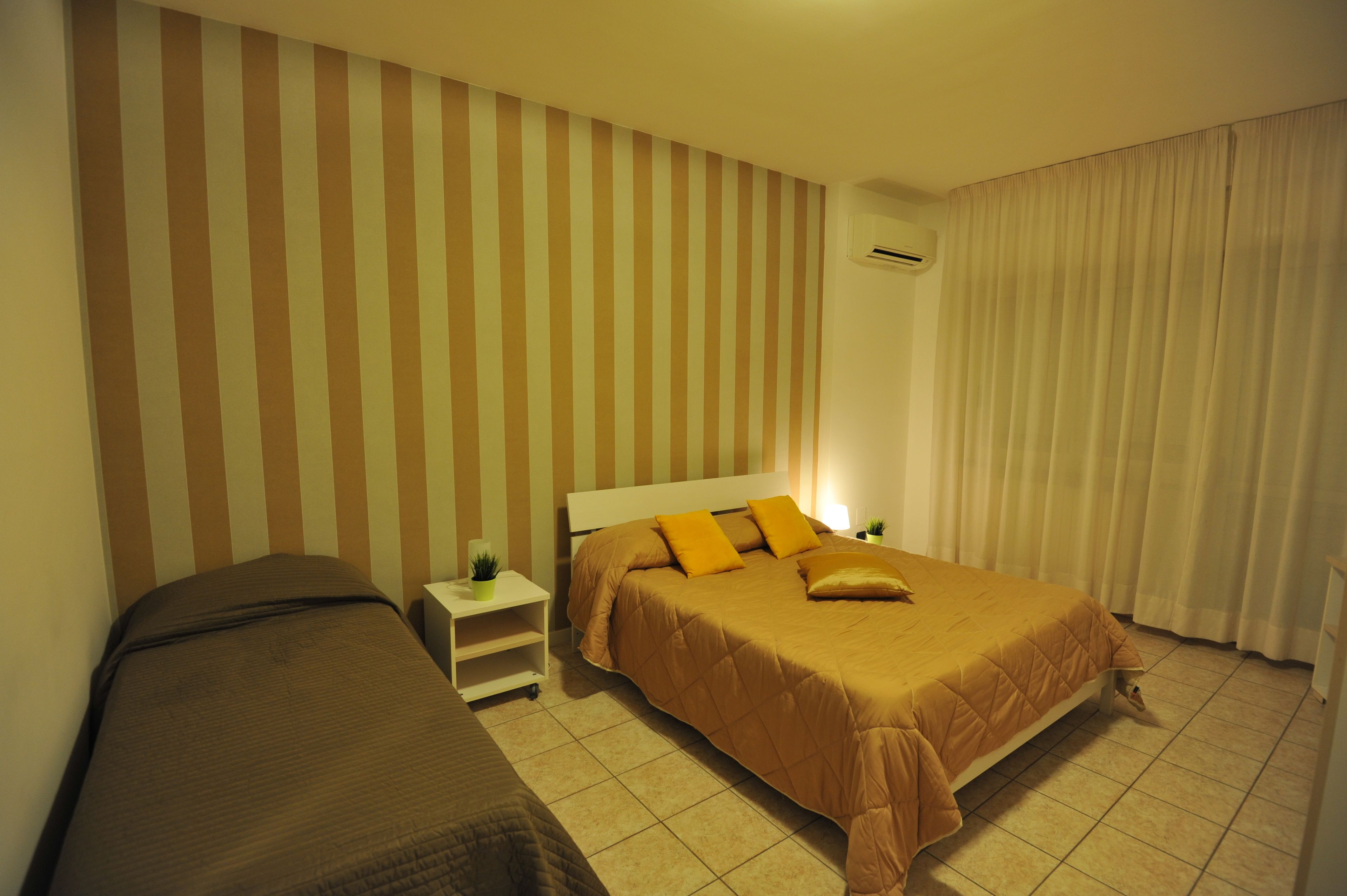 Una habitación de hotel con una pared a rayas, una cama con cubrecama marr ón, una mesita de noche con una planta y una ventana con cortinas.
