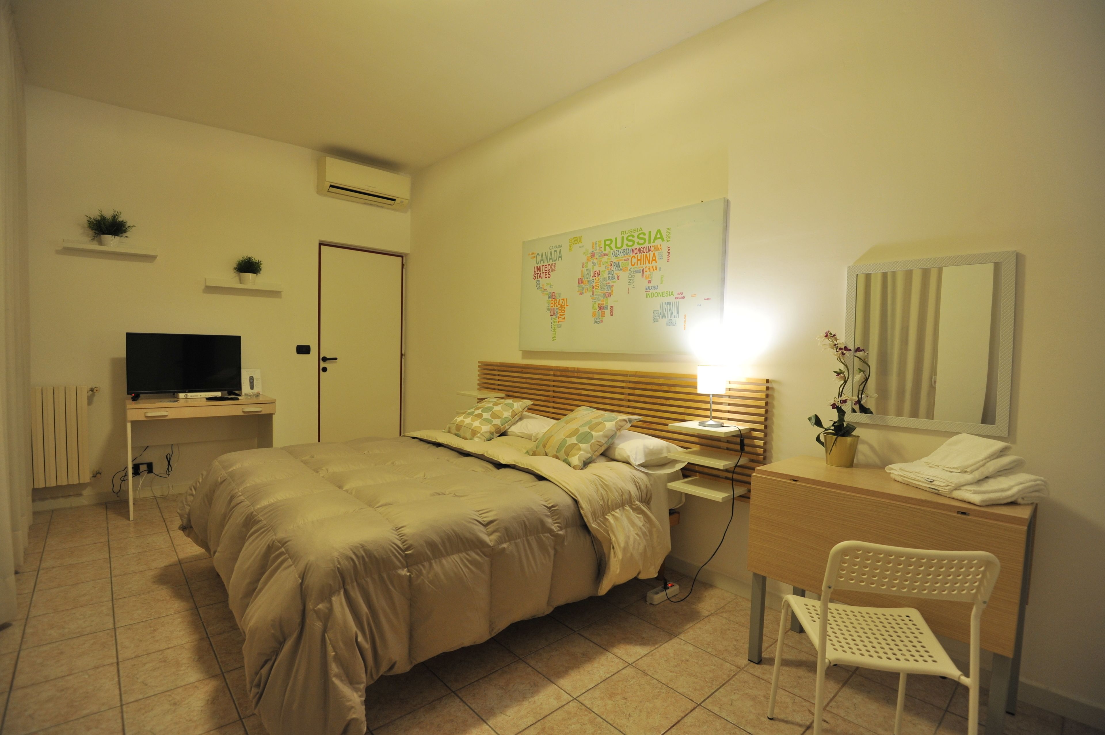 A bedroom with a bed, a desk, a chair, a TV, a map on the wall, and a mirror.