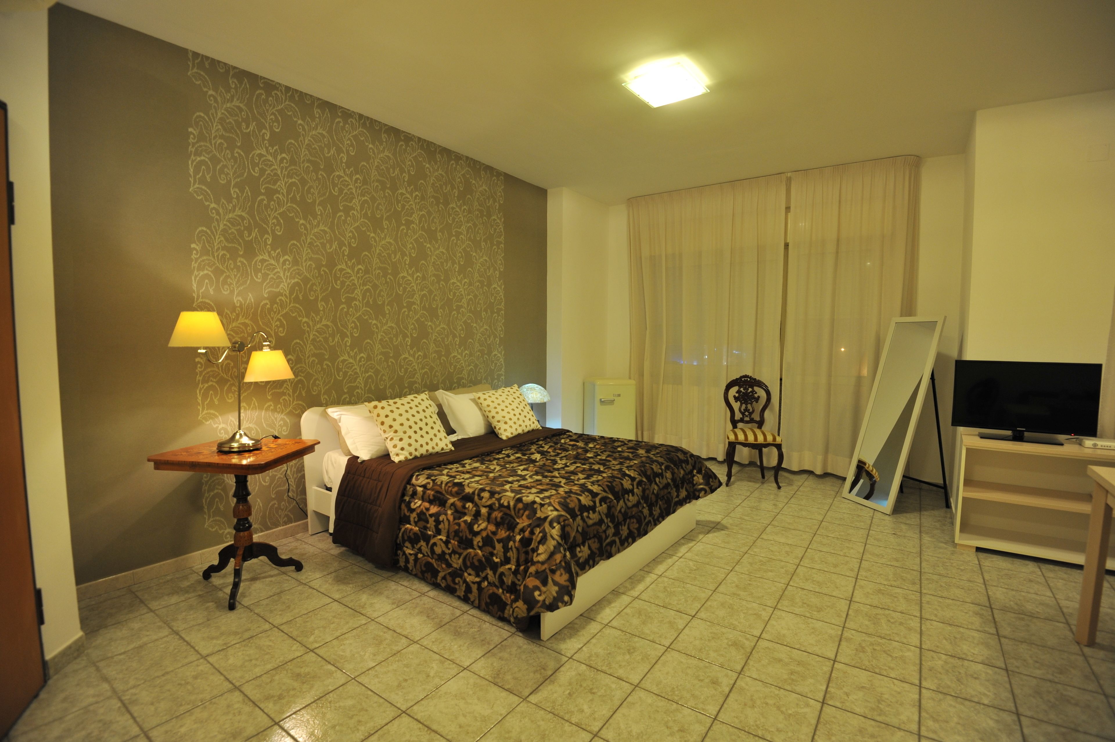 Habitación de hotel con cama, escritorio con lámpara, una silla, televisión y un gran ventanal con cortinas.