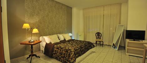 Habitación de hotel con cama, escritorio con lámpara, una silla, televisión y un gran ventanal con cortinas.