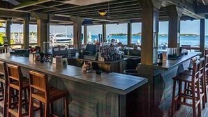 Una zona de bar con sillas de madera y una barra larga, con vistas a un cuerpo de agua con barcos.