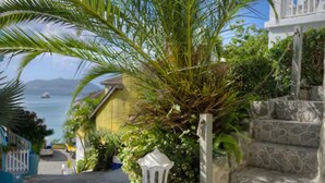 A coastal street with a stone staircase leading to a yellow building, surrounded by lush greenery and a view of the sea. - The Lighthouse Villas (Cane Garden Bay)