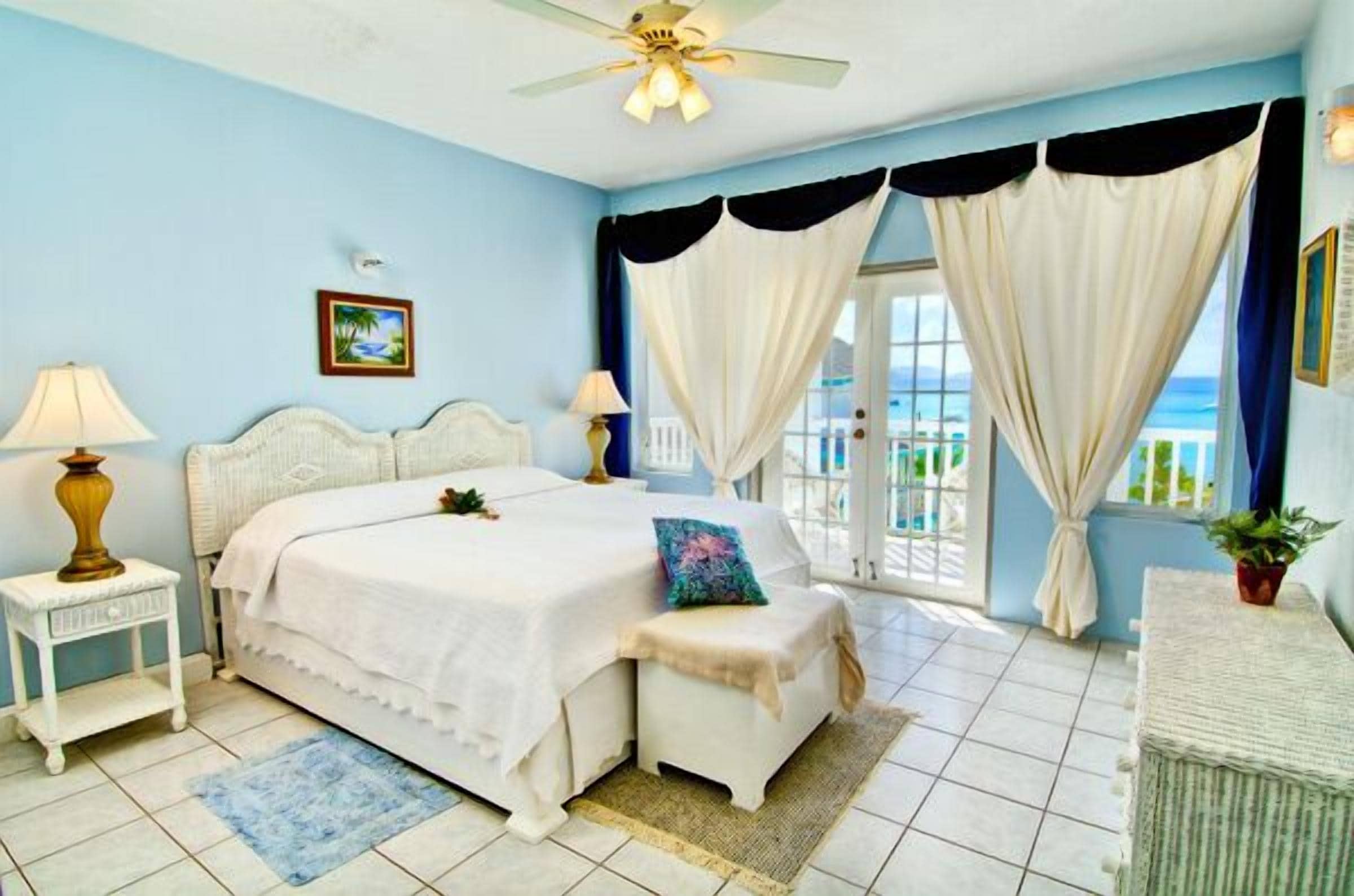 A bedroom with a bed, bedside tables, a bench, curtains, and a view of the outdoors.