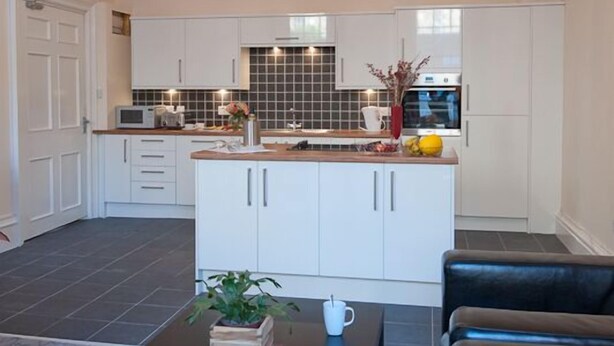 A modern kitchen with white cabinets, a central island, and a chandelier. There is a dining area with a table and chairs. - Windsor Street Apartments (Edinburgh)
