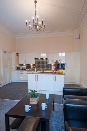 A modern kitchen with white cabinets, a central island, and a chandelier. There is a dining area with a table and chairs. - Windsor Street Apartments (Edinburgh)