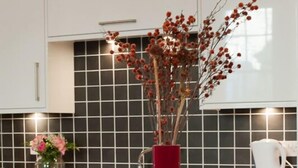 A kitchen with white cabinets, a black tiled backsplash, a red vase with dried branches, a glass bowl with a yellow squash, and a metal fruit basket. - Windsor Street Apartments (Edinburgh)