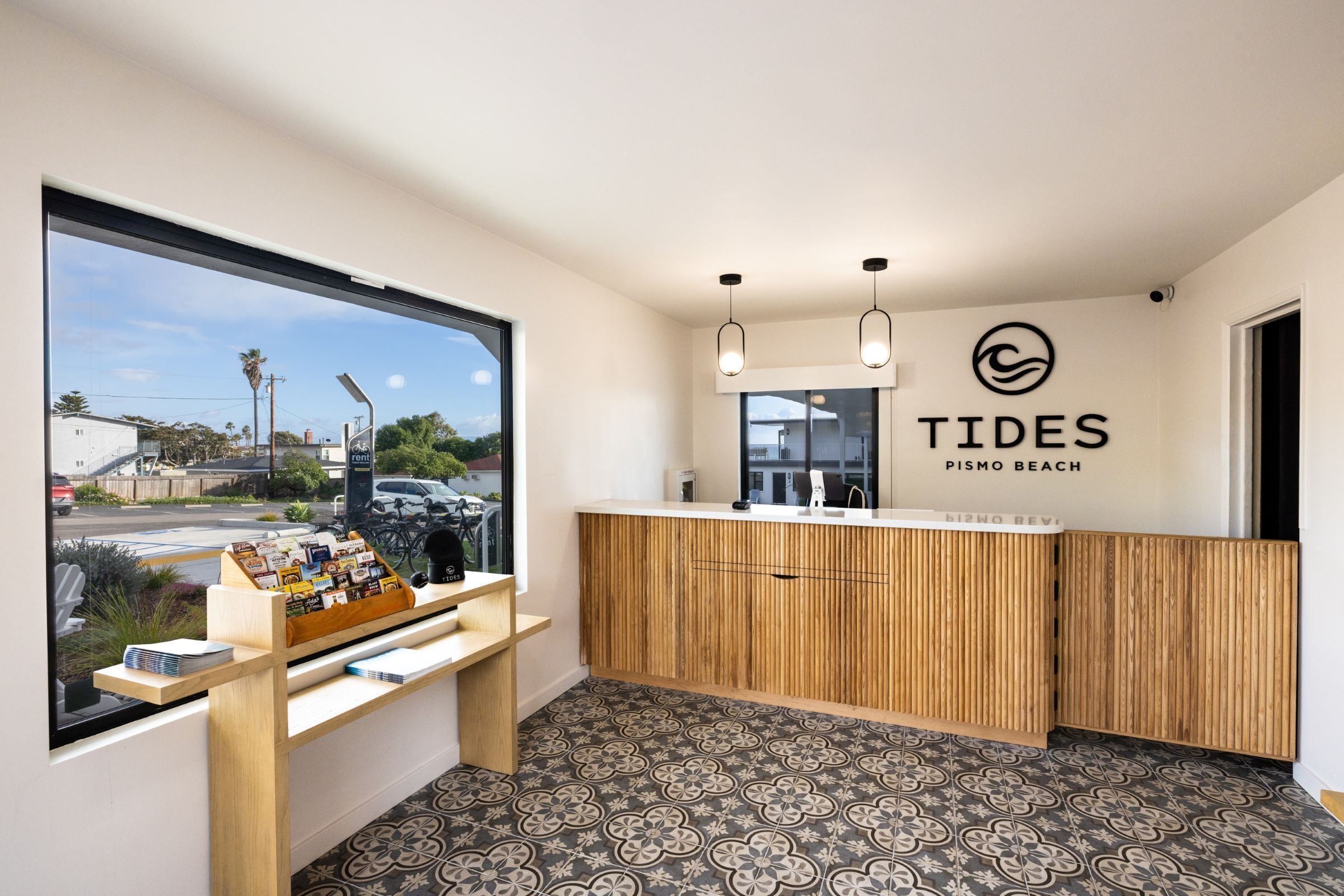 A reception area with a wooden counter, a large window offering a view of the street, and a wall sign reading "TIDES PISMO BEACH".