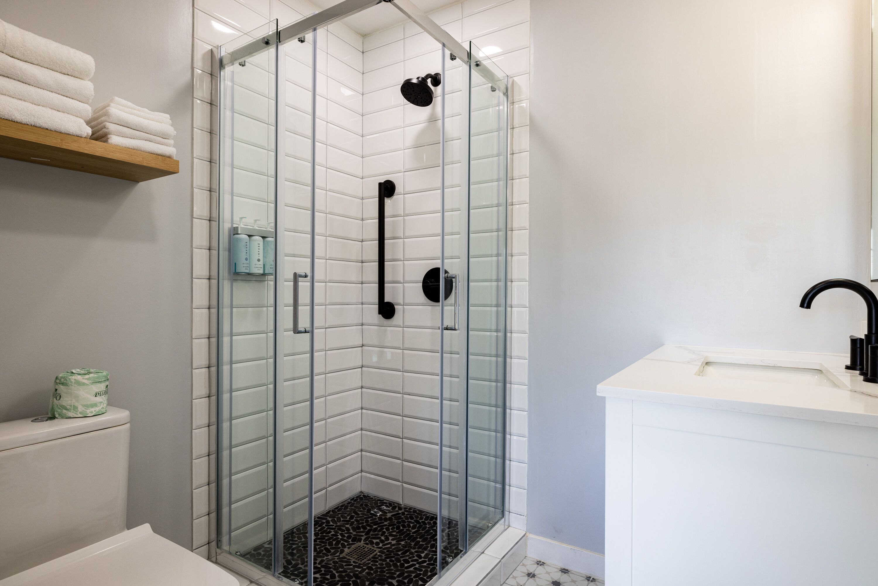 Een moderne badkamer met een douchecabine van glas, witte tegels, een witte wastafel en een houten plank met handdoeken.
