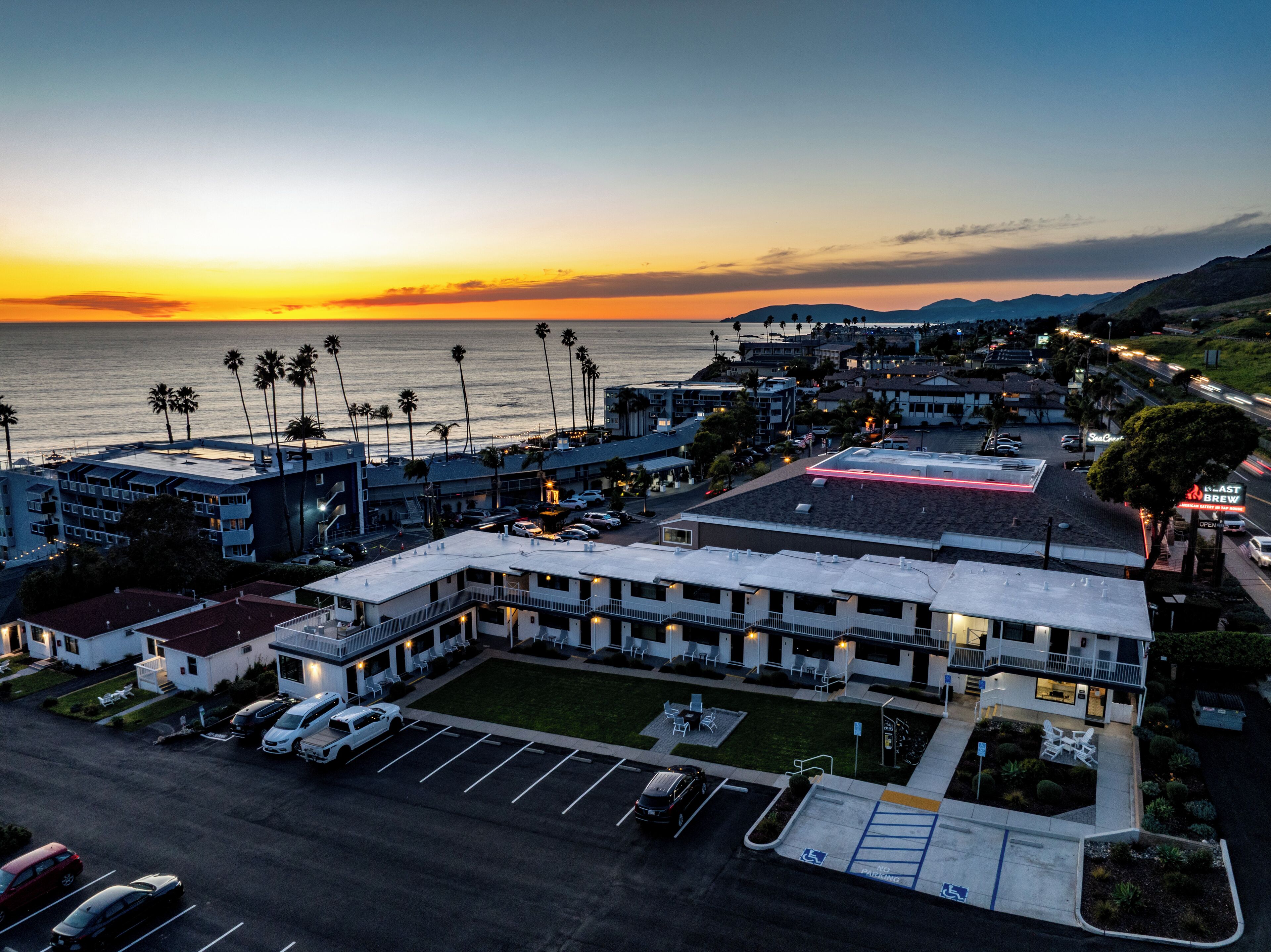 An aerial view of a coastal hotel with a parking lot, multiple buildings, and a view of the ocean at sunset.