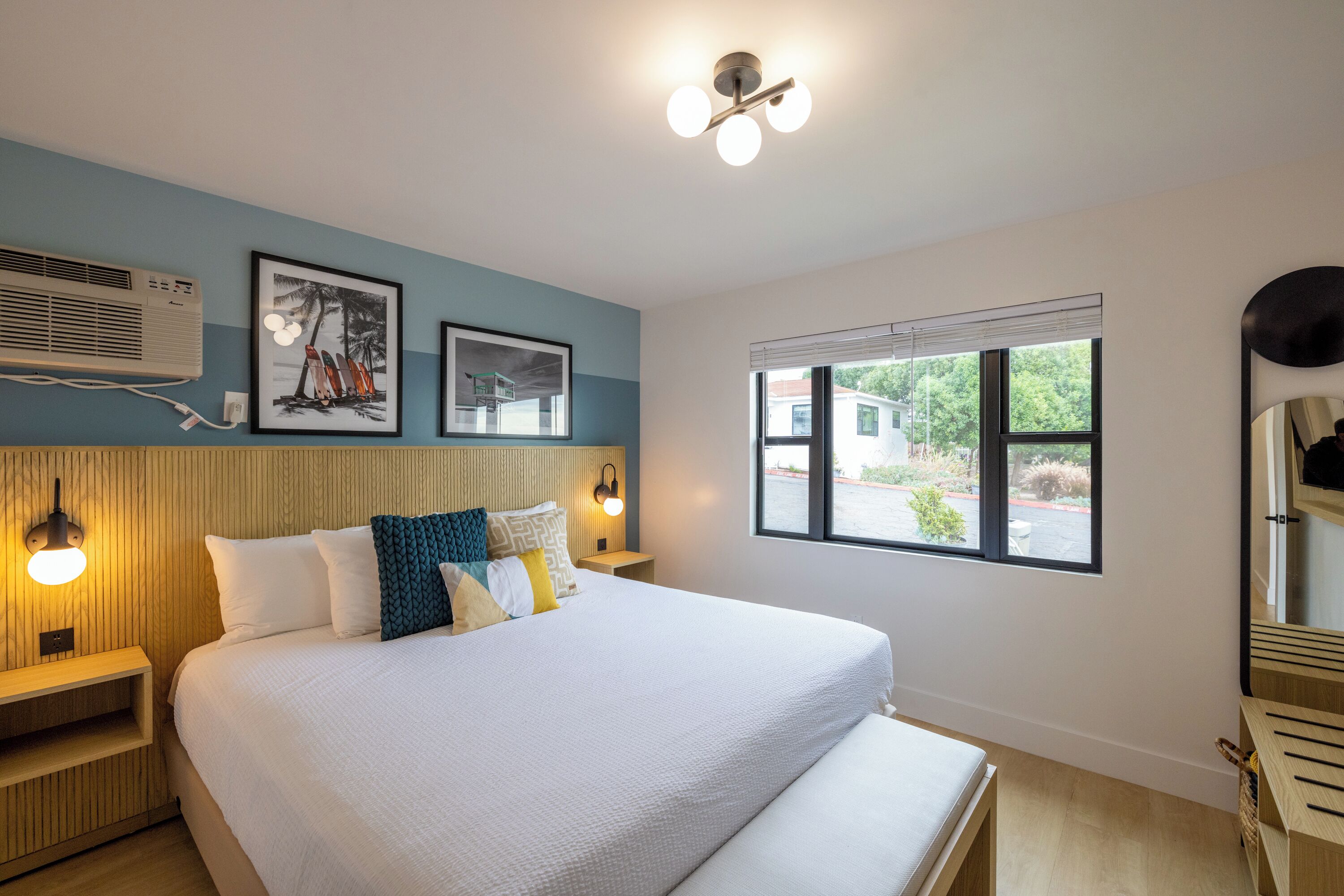 A modern hotel room with a large bed, a bedside table, a nightstand, a wall-mounted air conditioner, framed artwork, a window with a view, and a ceiling light fixture.
