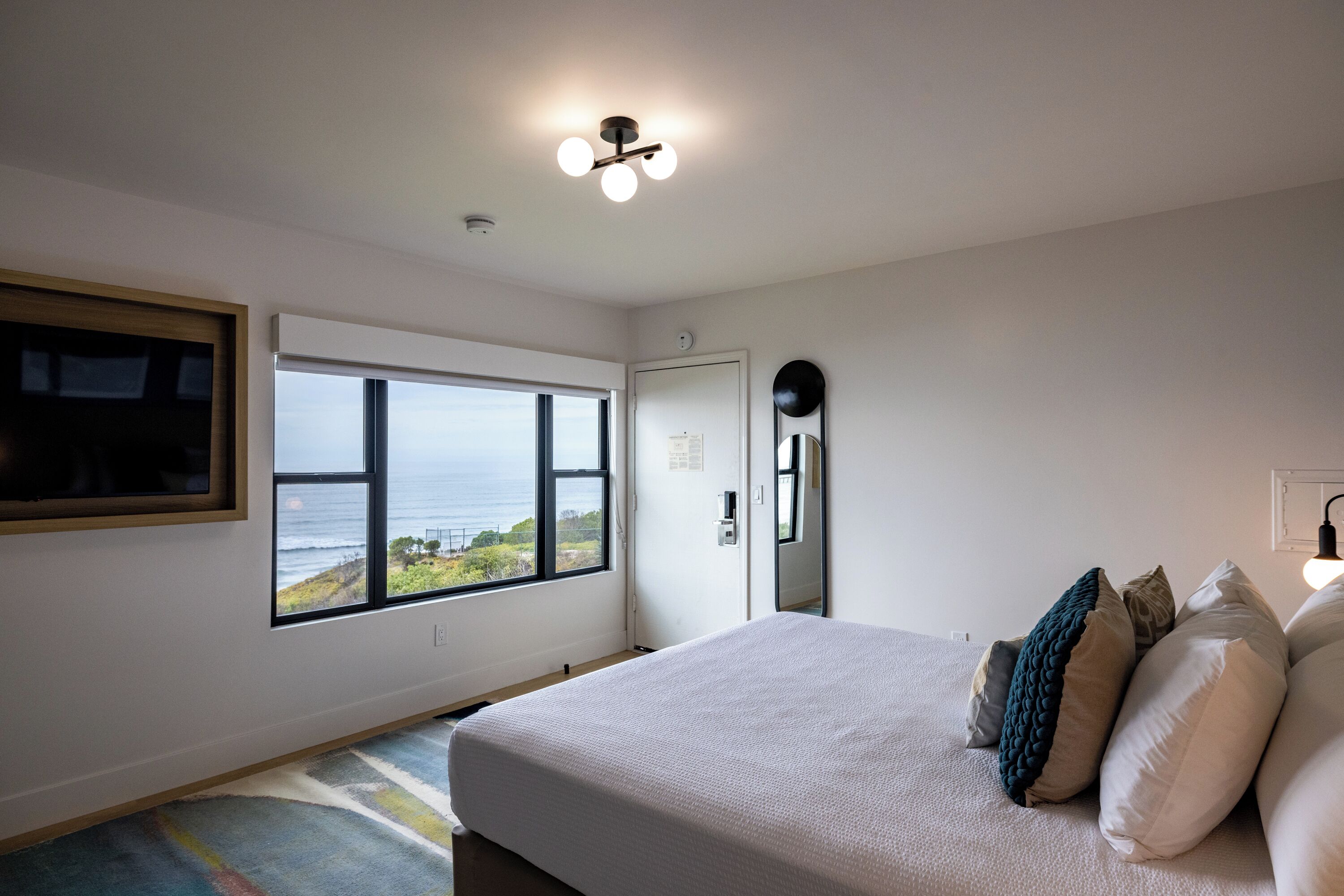 A bedroom with a large window, a bed with pillows, a ceiling light fixture, and a door.