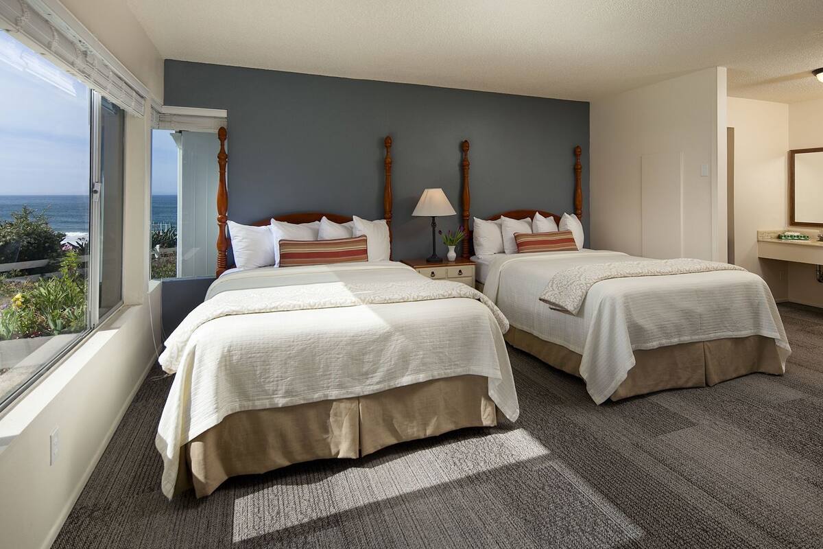 A hotel room with two beds, a nightstand, a lamp, a window with a view, and a bathroom with a mirror.
