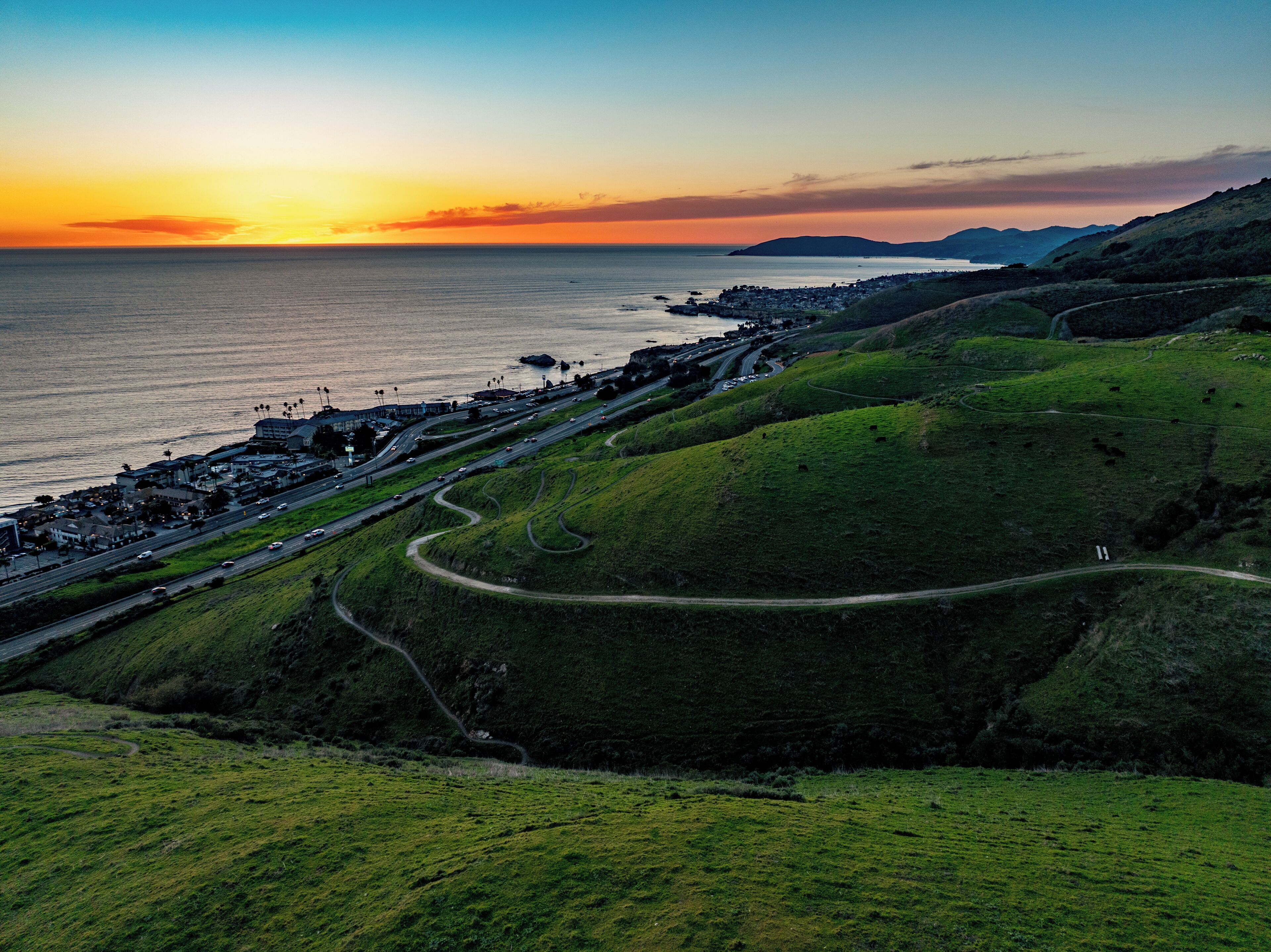 A coastal landscape with a winding road, green hills, and a sunset over the ocean.