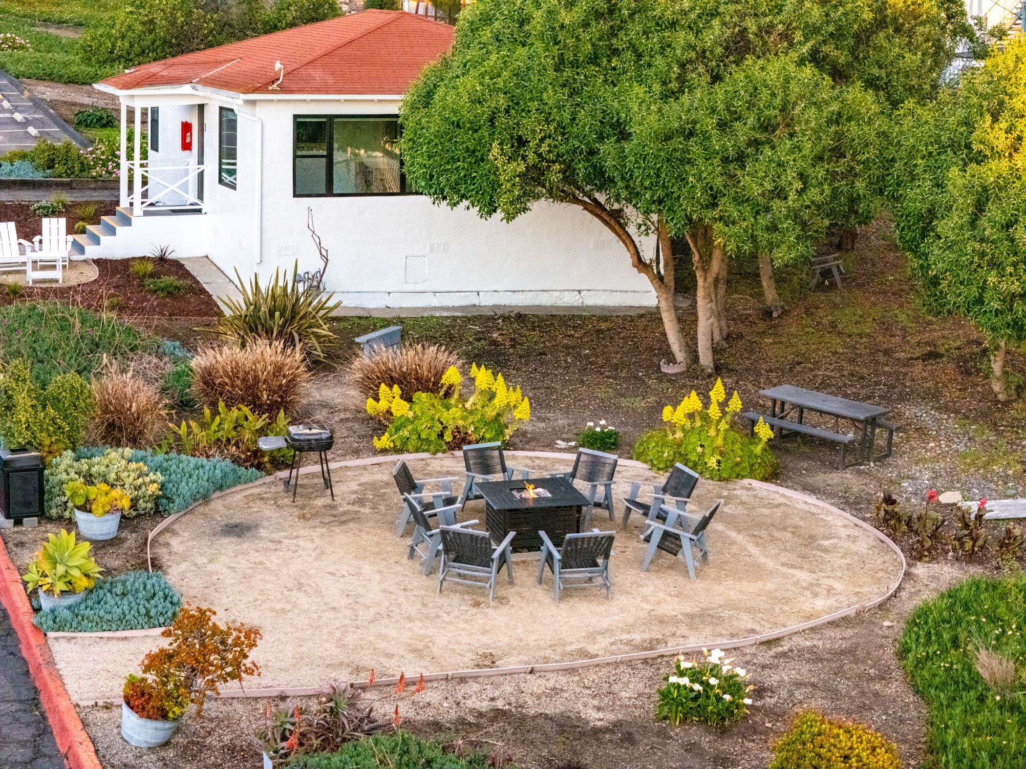 A patio area with a round table and chairs, surrounded by various plants and a white building in the background.