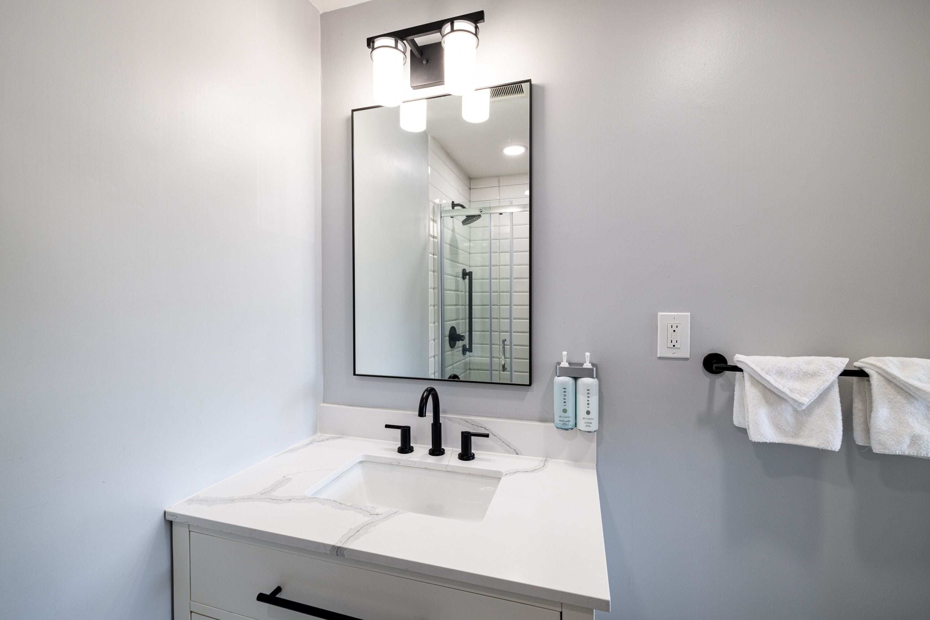 A bathroom with a white sink, black faucet, and a large mirror above it. There are two bottles of hand soap on the wall next to the mirror.