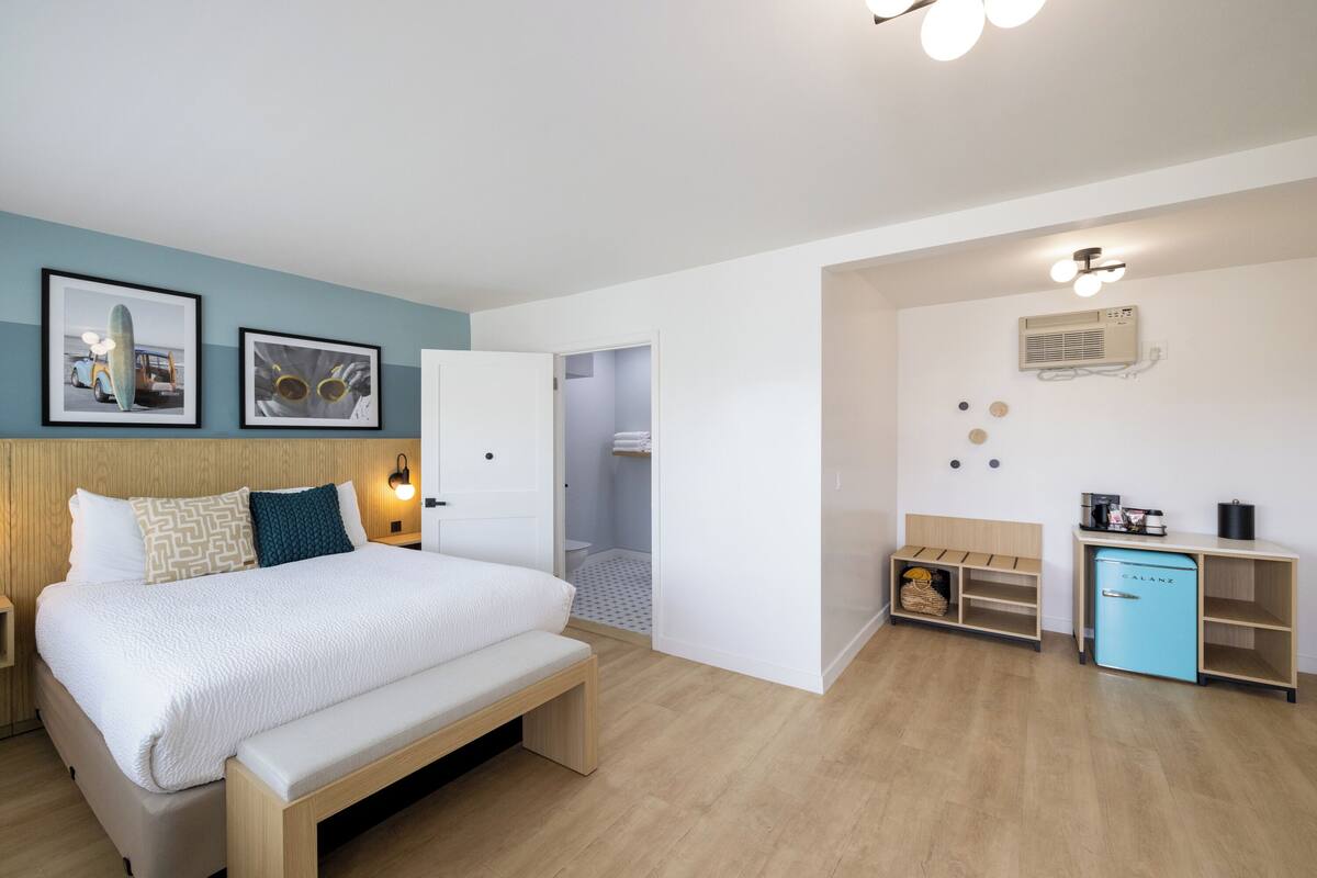 A modern bedroom with a bed, bedside tables, a bench, a wardrobe, a small kitchenette, and a bathroom.