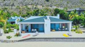 Una casa moderna frente a la playa con un porche cubierto, piscina y vistas de las colinas circundantes.