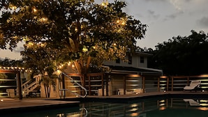 Un área de piscina con un árbol, luces colgantes, una escalera y sillas de descanso.
