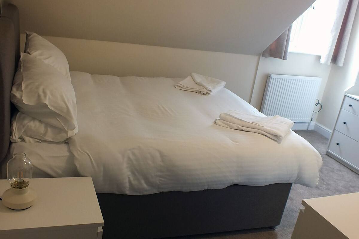A neatly made bed with pillows and a folded towel, a bedside table with a lamp, and a dresser with drawers.
