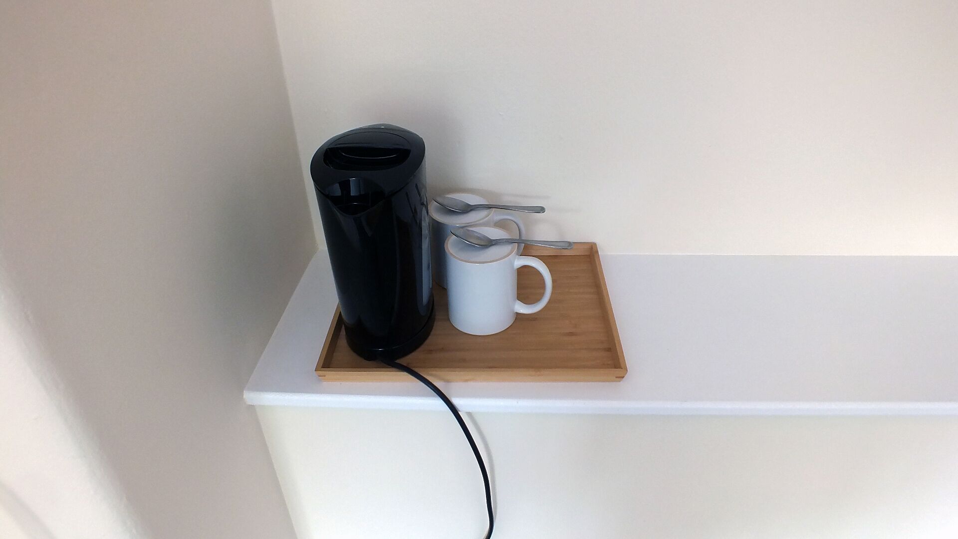 A black coffee maker on a white shelf with a white mug and a spoon on a wooden tray.