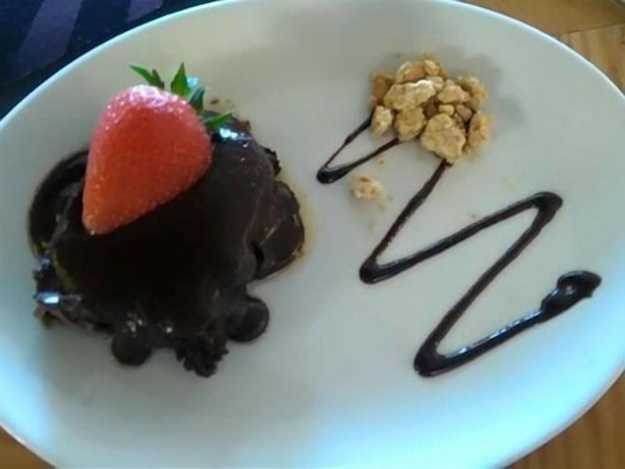 Une assiette de dessert avec un morceau de chocolat enrobé, surmonté d’une fraise et d’un petit tas de noix, le tout accompagné de quelques filets de sauce au chocolat.