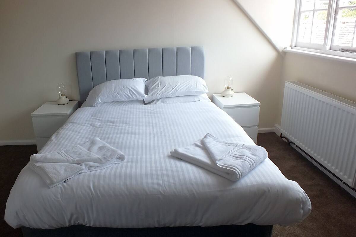 A neatly made bed with white linens and towels, a blue upholstered headboard, and white bedside tables with lamps.