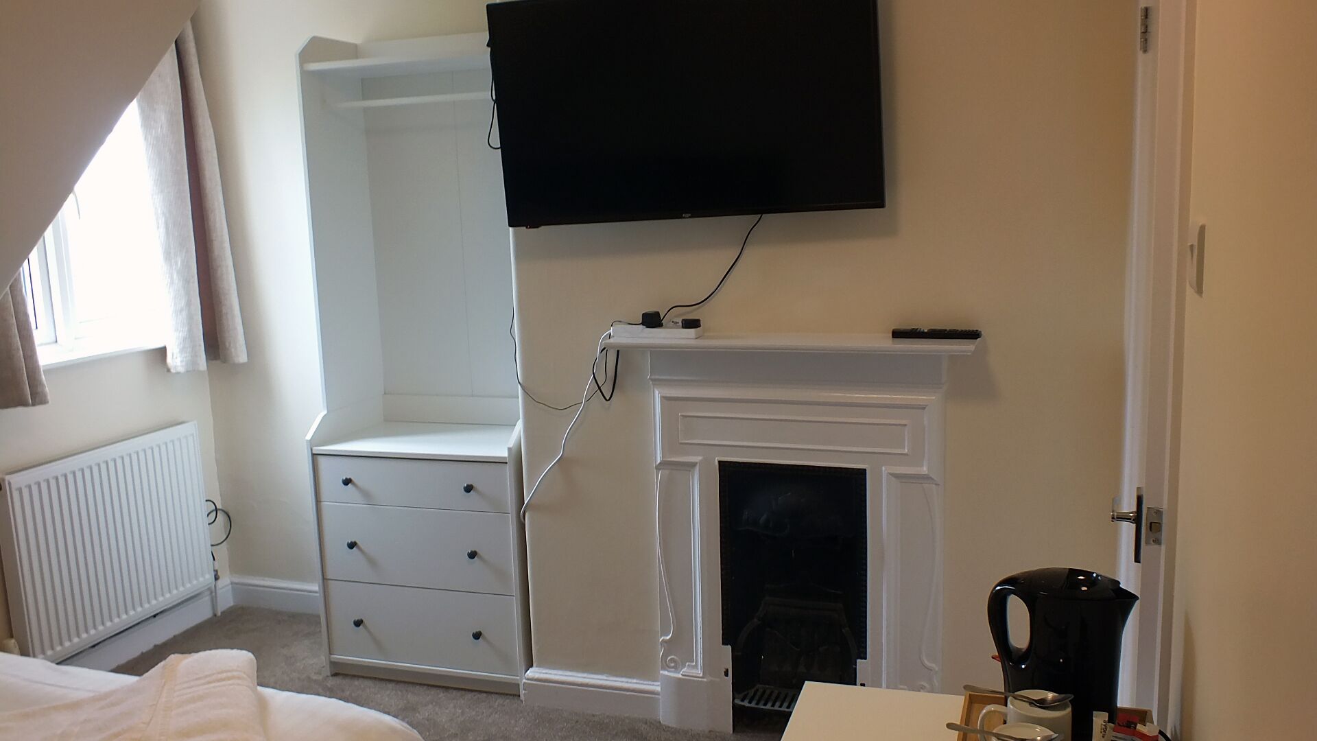 A bedroom with a white wardrobe, a fireplace, a television mounted above the fireplace, and a radiator.