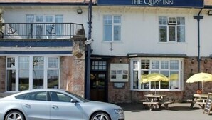 The Quay Inn, con un coche estacionado frente, mesas al aire libre con sombrillas amarillas y fachada de piedra.