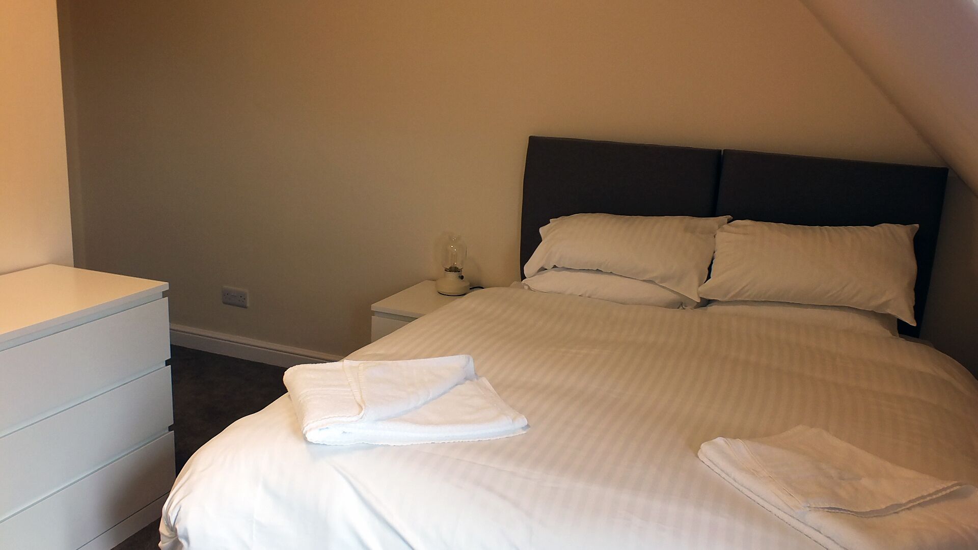 Una cama bien hecha con sábanas y almohadas blancas, una mesita de noche con lámpara y un aparador blanco.