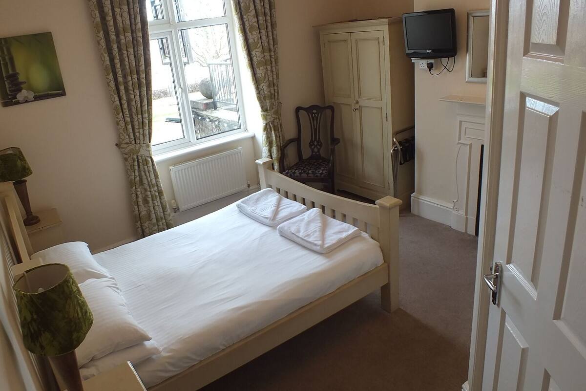 A small, clean bedroom with a single bed, a window with curtains, a television mounted on the wall, and a wardrobe.