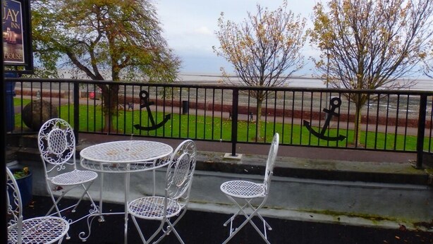 Un patio con sillas y mesas de metal blanco, una cerca de metal negro y vistas a un área verde con árboles y una masa de agua.